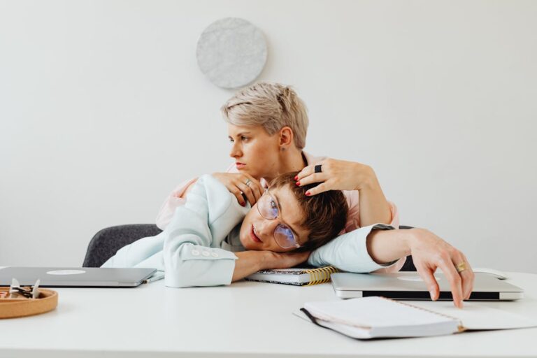 Two women leaning on each other at a desk, capturing a moment of work fatigue and support.