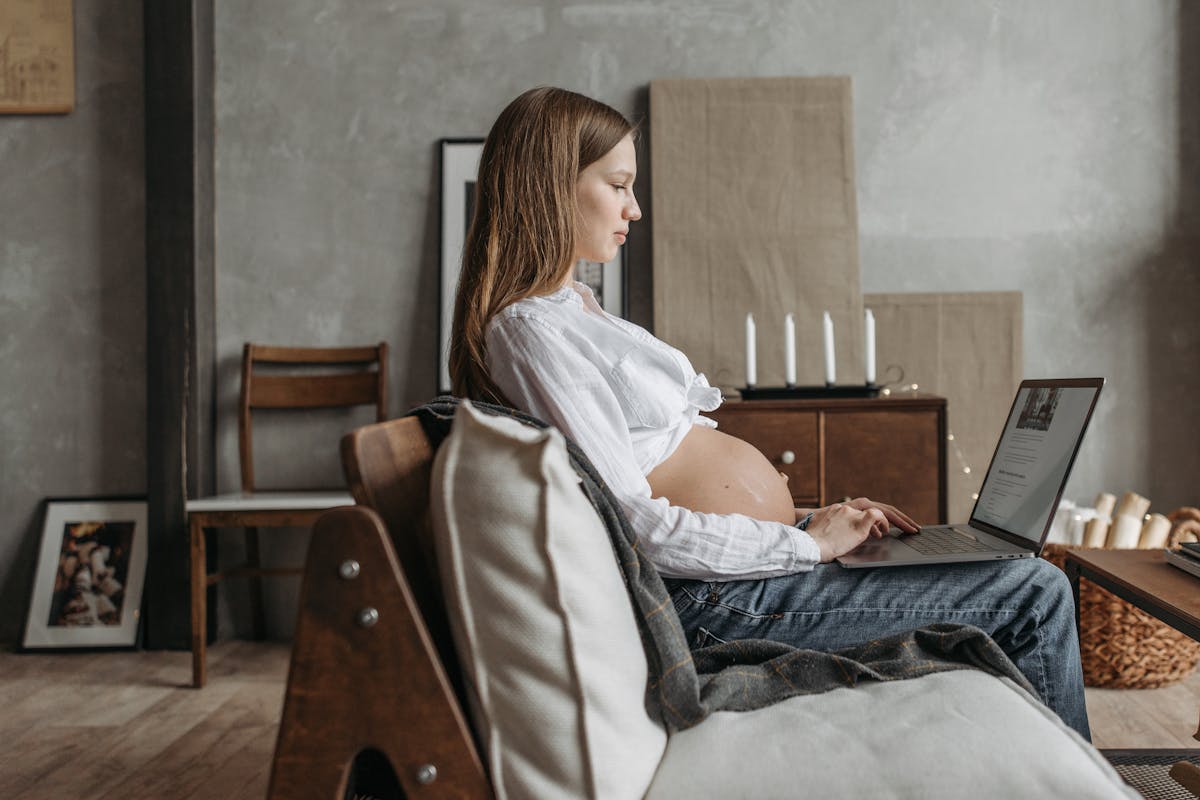 Side view of a pregnant woman using a laptop at home, emphasizing work from home lifestyle.