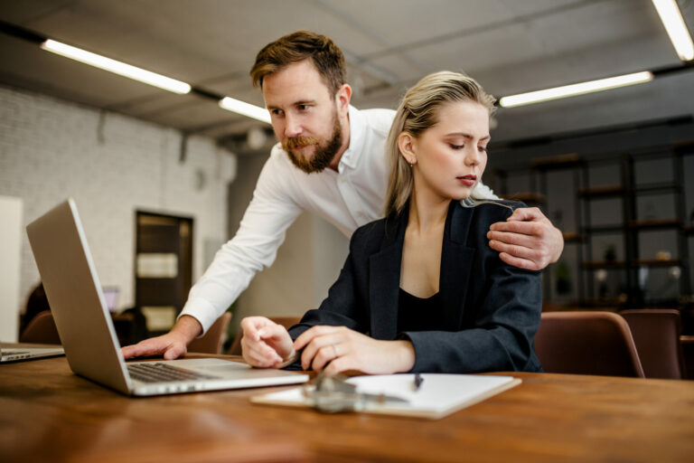 A bearded male Manager harasses his colleague a girl who works at a laptop in the office