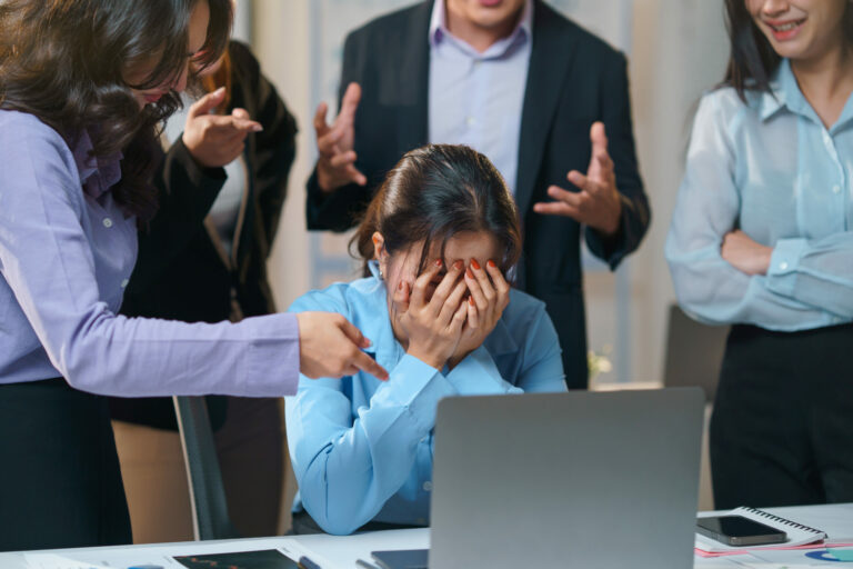 Colleagues pointing at businesswoman covering face with hands while sitting at desk in office meeting, concept of business failure and workplace bullying