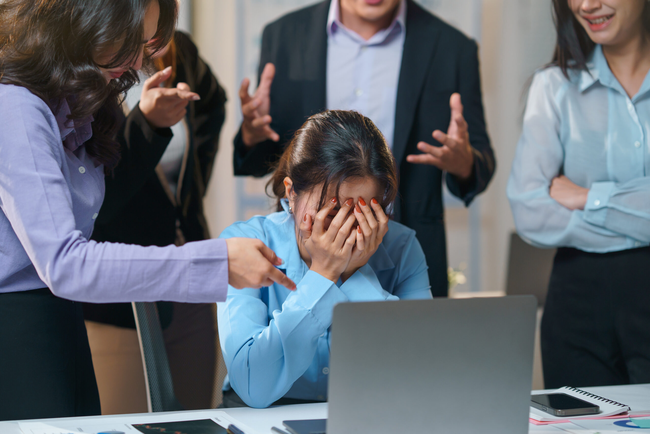 Colleagues pointing at businesswoman covering face with hands while sitting at desk in office meeting, concept of business failure and workplace bullying